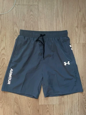 Grå träningsshorts från Under Armour - Snygga grå träningsshorts från Under Armour med vitt logotryck och text på benet. Shortsen har elastisk midja med snörning och är tillverkade i ett lätt och snabbtorkande syntetmaterial. Perfekta för gymmet eller löprundan. passar M