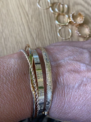 Mixade  armband och ringar - Säljer ett set med fyra armband i guld- och silverfärg. Armbanden har olika mönster, bland annat tvinnad design och graverade detaljer. De är stela bangles i metall och passar perfekt för dig som gillar att mixa stilar och skapa en personlig look. Samt åtta ringar i olika utfprmanden 