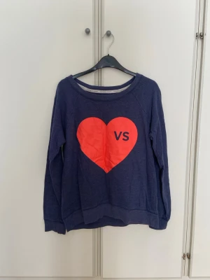 Blå sweatshirt med rött hjärta VS - Blå sweatshirt från Victoria's Secret med ett stort rött hjärta och VS-tryck på bröstet. Tröjan har rund halsringning, långa ärmar med ribbade muddar.