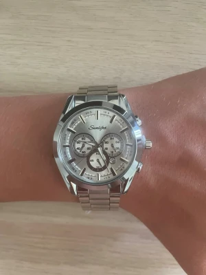 Silverfärgad klocka - Stilren klocka i silverfärgat metallarmband och boett. Urtavlan är också silver med flera mindre urtavlor och tydliga markeringar. Klockan har en klassisk och modern design som passar till många stilar.