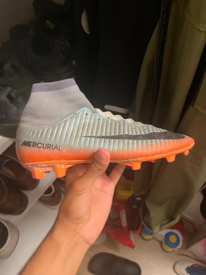 Nike Mercurial fotbollsskor grå/orange - Säljer ett par Nike Mercurial fotbollsskor med hög strumpkrage. Skorna är grå med orange sula och detaljer, samt svart Nike-logga på sidan. Ovandelen är i syntetmaterial med ribbad struktur för bättre bollkontroll. Perfekta för dig som vill ha snabbhet och stil på planen.
