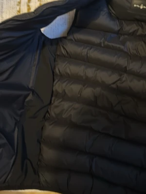 Svart dunväst från Sail Racing - Snygg svart dunväst från Sail Racing med quiltad design och dragkedja framtill. Västen har två praktiska fickor med dragkedja och logga på bröstet. Perfekt för lager-på-lager och håller dig varm med sin fluffiga dunfyllning.