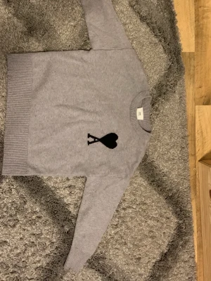 Grå ami paris tröja - Snygg grå stickad tröja med ett svart hjärta och bokstaven A på bröstet. Tröjan har rund hals, långa ärmar och breda ribbade muddar vid ärmslut och nederkant. Perfekt för dig som gillar stilrena detaljer och mjuka material.
