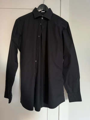 Svart klassisk skjorta - Stilren svart skjorta med lång ärm och klassisk krage. Skjortan har knappar framtill och är tillverkad i ett slätt tyg som ger en clean look. Perfekt för dig som gillar minimalistisk stil.