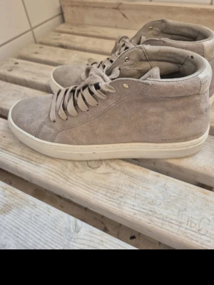 Beige mockasneakers med vita sulor - Snygga sneakers i beige mocka med vita sulor och matchande beige skosnören. Modellen är mellanhög och har en stilren design med diskreta sömmar och guldfärgade öljetter. Perfekta för en clean och avslappnad look.
