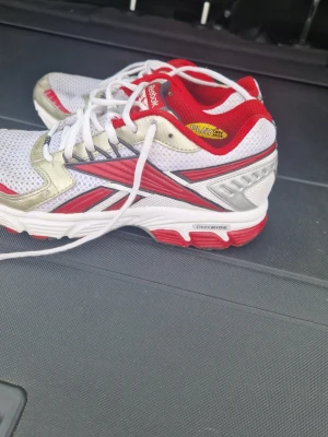 Vita och röda Reebok sneakers - Snygga Reebok sneakers i vitt och rött med detaljer i silver. Skorna har klassisk snörning och meshmaterial för bra ventilation. Ikonisk Reebok-logga på sidan och DMXRide-sula för extra komfort. Perfekta för dig som gillar sportig stil.