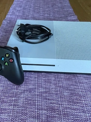 Xbox One S vit spelkonsol med kontroll - Säljer en Xbox One S spelkonsol i vit färg med en svart Xbox-kontroll och strömkabel. Konsolen har en stilren design med ventilerad ovansida och klassiska Xbox-knappar på kontrollen. Perfekt för gaming och streaming.