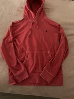 Röd/rosa hoodie från Polo Ralph Lauren - Snygg röd/rosa hoodie från Polo Ralph Lauren med klassisk huva och dragsnören. Har en känguruficka framtill och den ikoniska lilla loggan broderad på bröstet. Perfekt för en avslappnad och trendig stil. Den är washed Villket ger den en mer vintage look, står storlek xs men passar även S