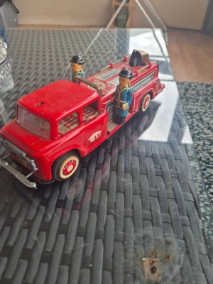 Retro röd plåtbrandbil med figurer - Säljer en retro leksaksbrandbil i röd plåt med detaljer i silver och vitt. Brandbilen har två brandmansfigurer i blå uniformer och svarta hjälmar. Modellen har stege, slang och klassiska dekaler på sidorna. Perfekt för dig som gillar samlarprylar eller vill ha en cool inredningsdetalj.