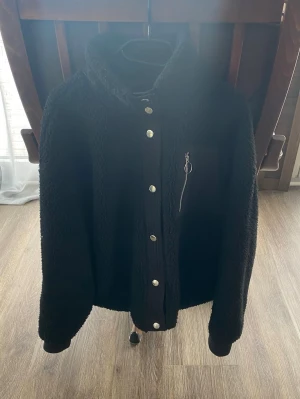 JDY Teddyjacka - stl XL - Mysig svart teddyjacka från JDY med huva och tryckknappar framtill. Jackan har en bröstficka med dragkedja och är gjord i fluffigt teddy-material som håller dig varm. Perfekt för dig som vill ha en stilren och bekväm look.