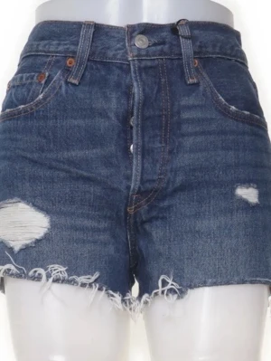 Levi's blå denimshorts med fransar - Snygga blå denimshorts från Levi's med slitna detaljer och fransig kant. Klassisk femficksmodell med hög midja och knappar framtill. Perfekta för sommaren och ger en cool, avslappnad vibe.