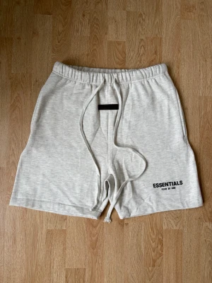 Grå Essentials shorts med snörning - Snygga ljusgrå shorts från Fear of God Essentials med elastisk midja och långa snören framtill. Klassisk relaxed fit och logga tryckt på vänster ben. Perfekta för chill dagar eller streetwear-stil. Tvär snygga och HELT nya aldrig använda eller tvättade, endast utan lapp men med original påse. Skriv gärna vid frågor eller funderingar.