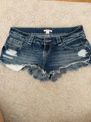 Blå slitna jeansshorts med fransar - Säljer ett par blå jeansshorts med slitningar och fransig kant. Shortsen har fem fickor och dekorativa sömmar på bakfickorna. De är tillverkade i denim och har en cool, avslappnad look.