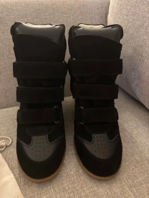 Isabel Marant skor Bekett  - Snygga svarta sneakers med högt skaft och flera breda kardborreband. Skorna har en mix av mocka och läderdetaljer samt en brun gummisula. Insidan är ljus och vadderad för extra komfort. Perfekta för dig som gillar streetstyle och vill sticka ut. Har kvitto från köpet (skriv privat) Box och påse medföljer!
