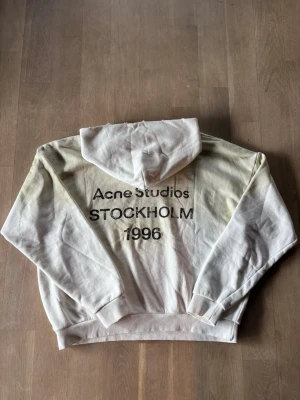 Vit hoodie Acne Studios Stockholm - Säljer en vit hoodie från Acne Studios med trycket 'Acne Studios STOCKHOLM 1996' på ryggen. Hoodien har en klassisk passform med huva och ribbade muddar vid ärmar och midja. Perfekt för dig som gillar minimalistisk och stilren streetwear.
