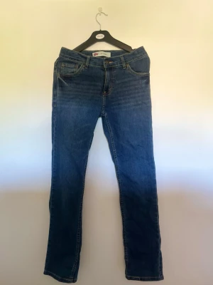Blå slim fit jeans från Levi's - Klassiska blå jeans från Levi's med rak passform och fem fickor. Jeansen har normal midja, dragkedjegylf och Levi's ikoniska läderpatch bak i midjan. Tillverkade i slitstark denim med snygga kontrastsömmar. Perfekta till vardagslooken.