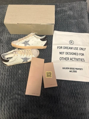 Golden Goose Superstar sneakers glitter - Golden Goose Superstar sneakers i vitt läder med silverglittrig stjärna på sidan och svartglittrig häl. Beige snörning och sula med vintage-look. Klassisk låg modell med coola detaljer och text på sidan.