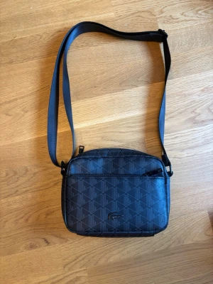 Svart axelväska från Lacoste - Snygg svart axelväska från Lacoste med justerbar rem och diskret mönster av Lacoste-loggor. Väskan har både fram- och bakficka med dragkedja och är tillverkad i slitstarkt syntetmaterial. Perfekt för att bära dina viktigaste grejer med stil.