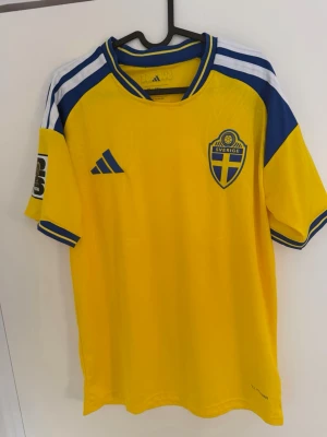 Svensk landslagströja Adidas gul - Säljer en gul svensk landslagströja från Adidas med blå detaljer och vita ränder på axlarna. Tröjan har korta ärmar, broderat Sverige-emblem på bröstet och turneringspatch på ärmarna. Perfekt för fotbollsfans som vill visa sitt stöd.