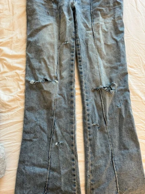 Ljusblå slitna bootcut jeans - Säljer ett par ljusblå bootcut jeans med råa sömmar och flera slitningar längs benen som jag har gjort själv. Jeansen har klassisk femficksdesign och dragkedjegylf. Perfekta för en avslappnad och trendig look.