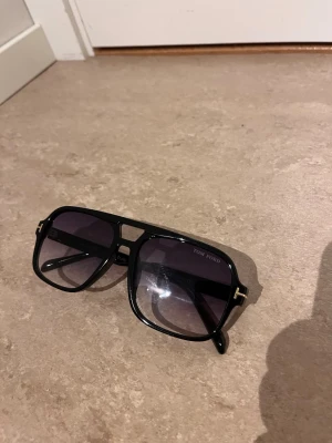 Svarta solglasögon från Tom Ford - Snygga svarta solglasögon från Tom Ford med klassisk pilotform och mörka tonade glas. Ramen är i blank svart plast med gulddetaljer vid gångjärnen. Perfekta för dig som vill ha en stilren och exklusiv look.