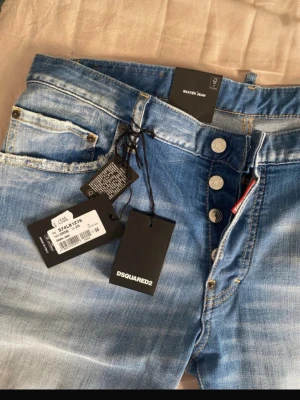 Blå Dsquared2 jeans med knappar - Säljer ett par snygga blå jeans från Dsquared2 med slitna detaljer och knäppning framtill. Jeansen har en klassisk femficksmodell och en tvättad look som ger en cool och avslappnad stil. Perfekta för dig som gillar streetwear och trendiga outfits.