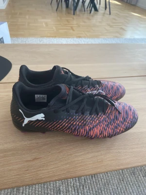 Puma fotbollsskor svart/lila/orange - Säljer ett par snygga Puma fotbollsskor med svart bas och ett mönster i lila och orange. Skorna har klassisk låg profil, snörning och greppvänliga dobbar i lila. Perfekta för dig som vill sticka ut på planen och få bra fäste. Tillverkade i syntetmaterial. Använda 1 gång 