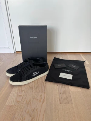 Svarta Saint Laurent sneakers - Svarta sneakers från Saint Laurent med vit logga på sidan och vit sula. Skorna har klassisk låg profil, snörning och är tillverkade i mocka. Medföljer originalkartong, dustbag och äkthetskort.