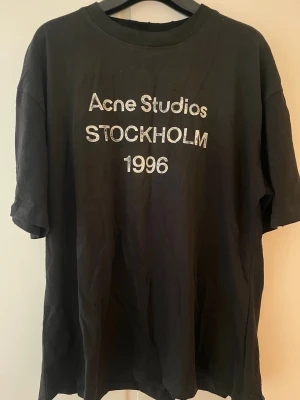 Svart Acne Studios t-shirt - Cool svart t-shirt från Acne Studios med tryck 'Acne Studios STOCKHOLM 1996' på bröstet. Oversized passform och klassisk rund halsringning. Perfekt till jeans eller shorts för en avslappnad look.