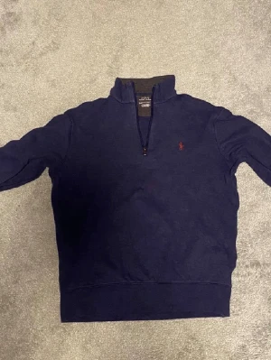 Marinblå halvzip från Polo Ralph Lauren - Snygg marinblå tröja med halv dragkedja från Polo Ralph Lauren. Klassisk design med hög krage och röd broderad logga på bröstet. Perfekt för lager-på-lager och chill stil. Mjuk bomullsblandning som känns skön mot huden.