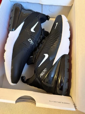 Nike Air Max 270 svarta sneakers - Säljer ett par Nike Air Max 270 sneakers i svart med vita detaljer och stor synlig Air-enhet i hälen. Ovandelen är i mesh och syntetmaterial för bra andningsförmåga. Snygg vit sula och logga på sidan. Perfekta för dig som gillar streetwear och sportig stil.