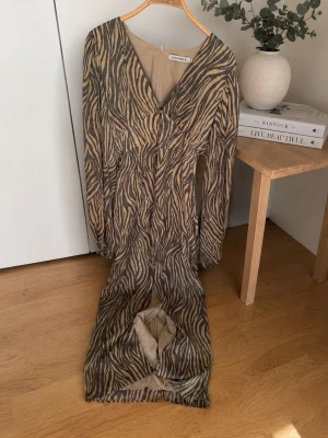 Zebramönstrad långklänning från Adoore - Snygg långklänning från Adoore med zebramönster i beige och svart. Klänningen har v-ringning, långa ärmar och en figurnära passform. Tillverkad i ett mjukt, lätt material som ger en elegant siluett.