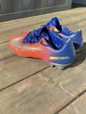 Nike Mercurial blå/röda fotbollsskor - Säljer ett par Nike Mercurial fotbollsskor i blått och rött med gula detaljer och silverfärgad swoosh. Skorna har snörning och är designade för spel på gräsplan. Lätta och smidiga med syntetiskt material för bra bollkontroll. Framdelen har släppt som visat på bild men går att spela i vanligt då det är ihoplimmat med ett starkare lim. ACC modellen vilket är den dyraste av Nike mercurial.
