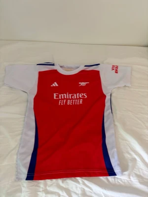 Arsenal matchtröja Saka #7 - Röd, vit och blå Arsenal matchtröja från Adidas med Saka #7 på ryggen. Tröjan har korta ärmar, klubbmärke och sponsortryck på bröstet samt detaljer i blått längs sidorna. Perfekt för dig som älskar fotboll och vill stötta Arsenal.