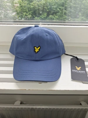 Blå keps från Lyle & Scott - Snygg blå keps från Lyle & Scott med klassisk gul broderad logga framtill. Kepsen har böjd skärm och justerbar rem bak. Tillverkad i mjuk bomull som känns skön att bära. Perfekt accessoar för en avslappnad och stilren look.