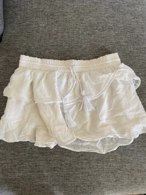 Vit volangkjol med snörning - Supersöt vit kortkjol med volanger och snörning i midjan med tofsar. Säljer då den inte passar längre💕 Ifrån Gina Tricot Young storlek 170