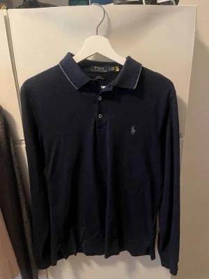 Marinblå långärmad skjorta Polo Ralph Lauren - Klassisk mörkblå långärmad pikétröja från Polo Ralph Lauren med krage, knäppning framtill. Inga slitage på tröjan.