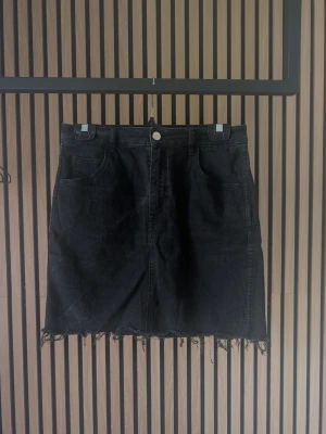 Svart jeanskjol med fransad kant - Säljer en svart jeanskjol med fransad nederkant och klassisk femficksdesign. Kjolen har normal passform och stängs med knapp och dragkedja framtill. Perfekt till en avslappnad och trendig look.