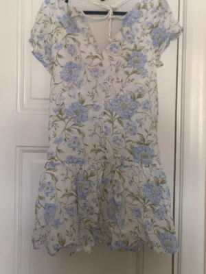 Blommig kortklänning med snörning - Söt kortklänning i vitt med blått blommönster och gröna blad. Klänningen har v-ringning med dekorativ snörning och korta ärmar. Volangdetalj nedtill ger en luftig känsla. Perfekt för varma dagar och sommarens alla tillfällen.