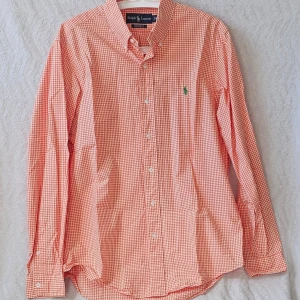 Rutig skjorta från Ralph Lauren orange - Klassisk orange och vit rutig skjorta från Ralph Lauren med button-down krage och grönt broderat logotyp på bröstet. Skjortan har långa ärmar, knappar framtill och är tillverkad i mjuk bomull. Perfekt för en avslappnad men ändå stilren look.