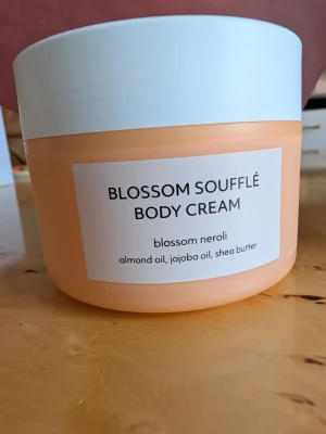 Blossom Soufflé Body Cream Estelle & Thild - Oöppnad Blossom Soufflé Body Cream från Estelle & Thild med doft av blossom neroli. Innehåller mandelolja, jojobaolja och sheasmör. Kommer i en rund, persikofärgad burk med vitt lock. Ekologisk och vegansk formula.