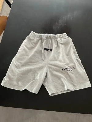 Grå Essentials shorts med snörning - Snygga ljusgrå shorts från Fear of God Essentials med svart logga på benet. Shortsen har elastisk midja med snörning och är tillverkade i mjuk bomull. Perfekta för en avslappnad och trendig look.