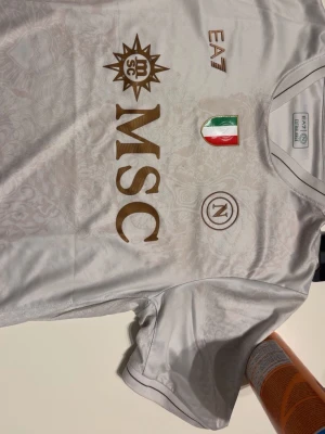 Vit Napoli fotbollströja EA7 - Snygg vit fotbollströja från Napoli med guldfärgade detaljer och loggor. Tröjan har MSC och EA7 tryck samt italienska flaggan på bröstet. Tillverkad i ett lätt och ventilerande material som passar perfekt till träning eller match.