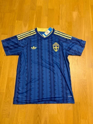 Sveriges vm tröja L - Säljer en blå fotbollströja från Adidas med Sveriges landslagsemblem på bröstet och gula detaljer. Tröjan har korta ärmar, rund hals och tre gula ränder på axlarna. Materialet är lätt och andas, perfekt för träning eller match.