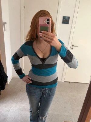 Randig långärmad topp från Ralph Lauren - Snygg långärmad topp från Ralph Lauren med breda ränder i turkos, grått och svart. Modellen har v-ringning med knappdetalj och figurnära passform. Perfekt till jeans för en avslappnad men ändå stilren look.