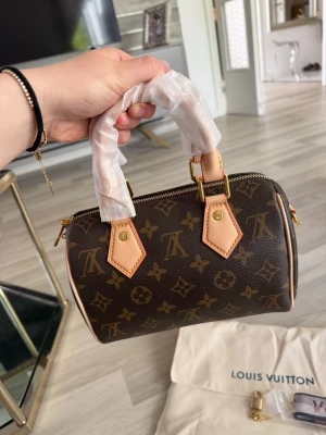 Väska  - Säljer en Speedy Nano i klassiskt brunt monogramcanvas med beige läderdetaljer och guldfärgade metalldelar. Väskan har två handtag, röd textilinsida och dragkedja. Dustbag, axelrem och lås medföljer. Perfekt för dig som gillar lyxiga accessoarer. Pris kan diskuteras vid snabb affär. Hör gärna av er vid frågor! 