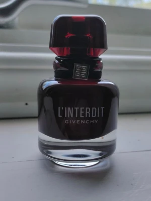 Givenchy L'Interdit parfymflaska - Givenchy L'Interdit parfym i en elegant, mörkröd glasflaska med genomskinlig botten och en stilren, fyrkantig kork. Flaskan har en silverfärgad Givenchy-logga runt halsen och texten L'INTERDIT på framsidan. Perfekt för dig som gillar lyxiga dofter.