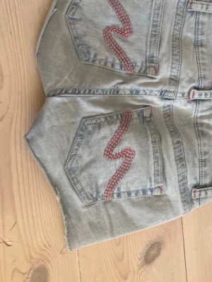 Ljusblå jeansshorts med broderi - Säljer ett par ljusblå jeansshorts med råa kanter och röda broderade detaljer på bakfickorna. Shortsen har klassisk femficksdesign och bälteshällor. Perfekta för varma dagar och en avslappnad stil. Storleken står de 40 men jag har 36 och de passar på mig 