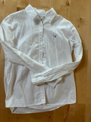 Vit skjorta från GANT med broderad logga - Klassisk vit skjorta från GANT med broderad logga på bröstet. Skjortan har knappar framtill, lång ärm och en tidlös design som passar till allt. Tillverkad i mjuk bomull för en bekväm känsla.