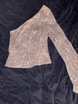 Glittrig one shoulder topp från NA-KD - Säljer en glittrig, beige one shoulder topp från NA-KD. Toppen är täckt av paljetter och har en lång ärm på ena sidan medan den andra axeln är bar. Perfekt för dig som vill sticka ut med en trendig och snygg look.